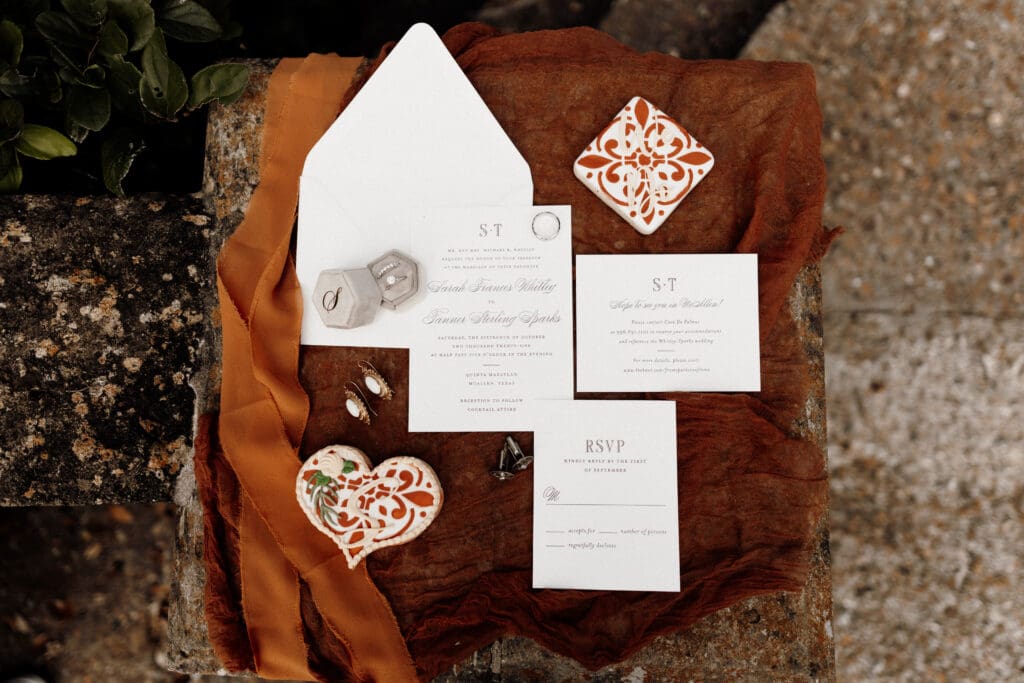 Wedding invitation suite with decorative elements on a textured surface. Includes an invitation, RSVP card, and an envelope. Accented with a brown cloth, ceramic ornament, rings, and ring box. Ideal for couples seeking the artistry of a San Antonio wedding photographer. Greenery visible in the corner.