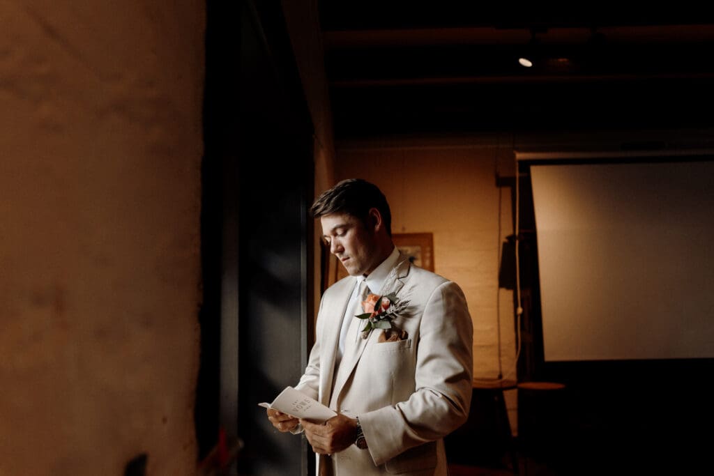A person in a white suit, adorned with a floral boutonniere, stands indoors reading from a paper. The room is dimly lit with a projector screen visible in the background, capturing the moment beautifully like an Edinburgh wedding photographer would at a memorable ceremony.