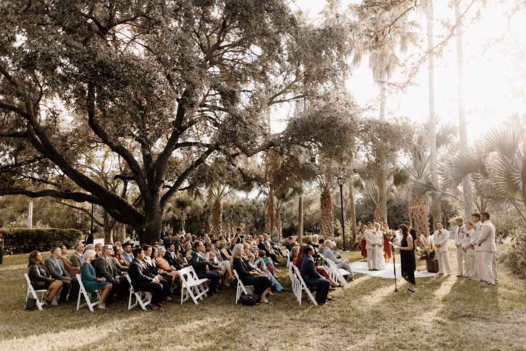 A wedding ceremony unfolds outdoors with guests seated on white chairs under a large tree. The wedding party stands to the right, wearing coordinated attire. Sunlight filters through the trees, casting a warm glow—perfect for any McAllen wedding photographer capturing this enchanting moment.