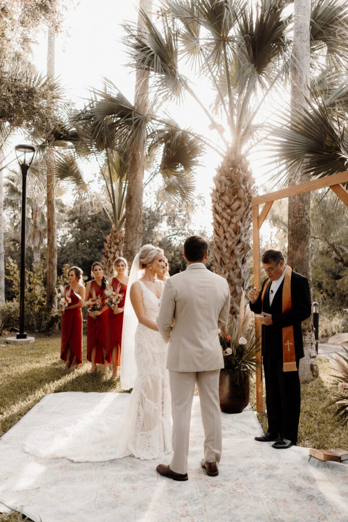 A couple stands at an outdoor wedding ceremony under tall palm trees, captured by a talented McAllen wedding photographer. The bride, in a lace gown and veil, faces the groom in a light suit. Bridesmaids in red dresses stand to the side while an officiant stands before a wooden arch.