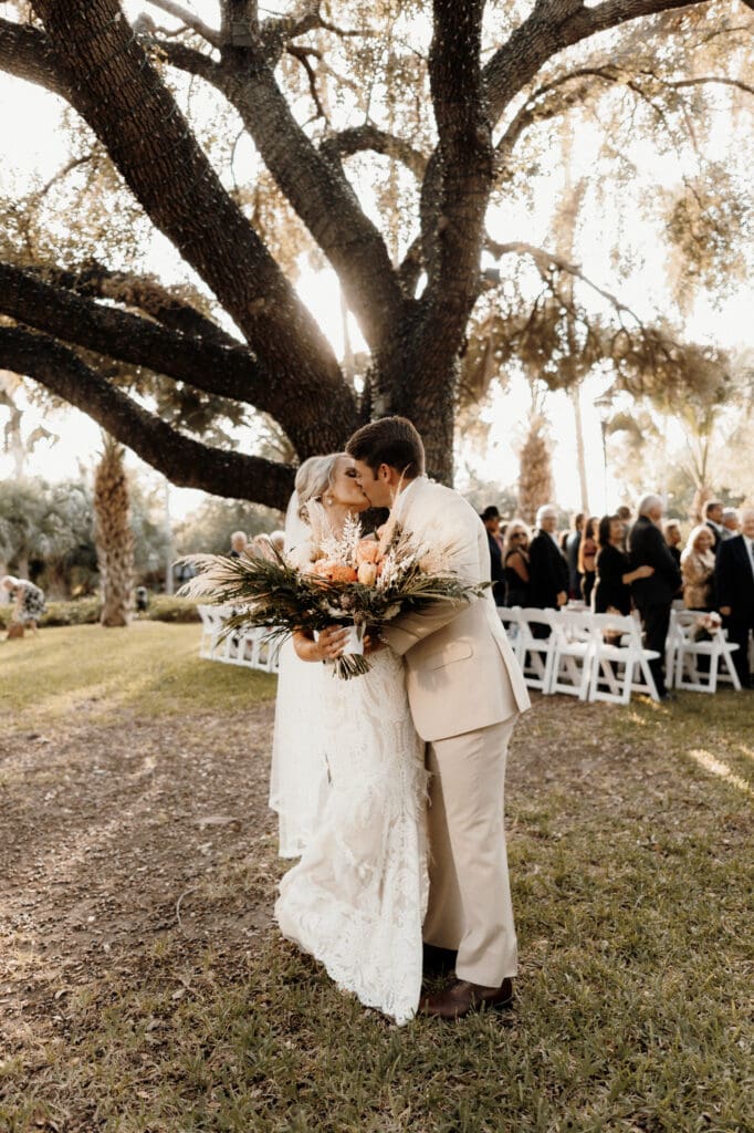 A bride and groom share a kiss under a large tree at an outdoor wedding ceremony, beautifully captured by a San Antonio wedding photographer. The bride holds a bouquet of flowers as guests are seated on white chairs in the sunlit and serene scene.