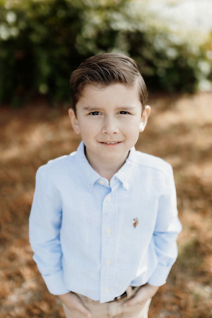 A young boy with short, dark hair stands outdoors on a sunny day. Dressed in a light blue button-up shirt and khaki pants, he stands amidst greenery. Captured by a talented McAllen wedding photographer, he looks directly at the camera with a neutral expression.