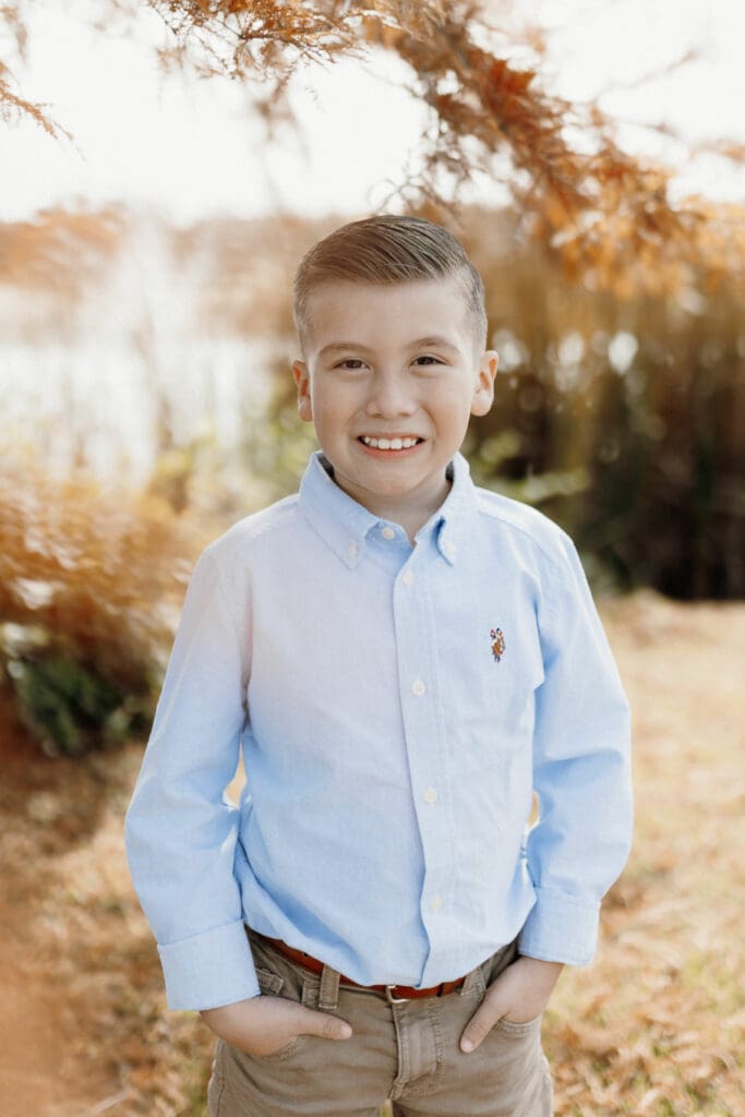 A young boy in a light blue button-up shirt smiles with his hands in his pockets. He stands outdoors in front of tall grass and autumn-toned foliage, a picturesque scene captured by a talented wedding photographer in McAllen.