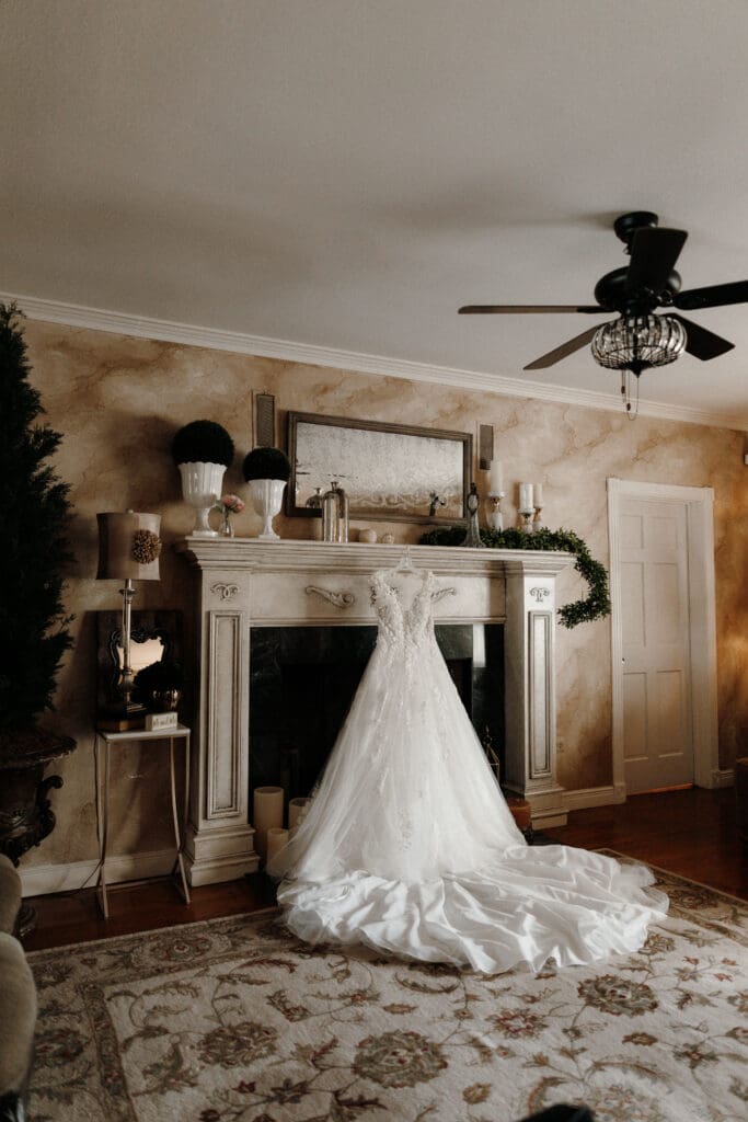 A wedding dress with a long train is draped over a fireplace mantle in an elegantly decorated room. Captured by a South Padre Island wedding photographer, the scene features a ceiling fan, potted plants, candles, and a large rug on the wooden floor.