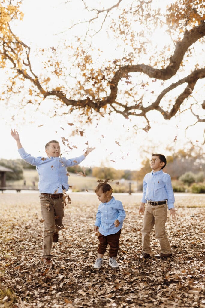 Three children wearing blue shirts and jeans playfully throw and kick autumn leaves in a park, much like a joyful scene captured by an Edinburg wedding photographer. The ground is covered with fallen leaves, leafless branches arch overhead, and the scene is filled with laughter and the warm colors of fall.