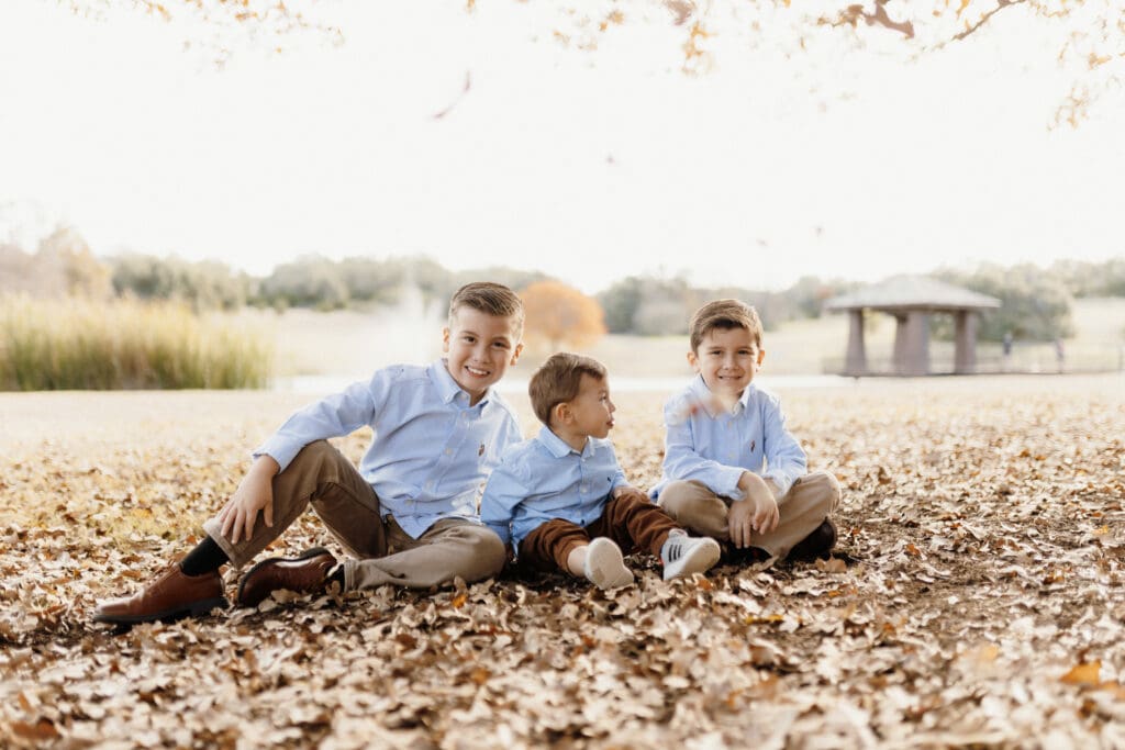 Three young boys sit on fallen leaves in a park, wearing light blue shirts and khaki pants. A grassy field, trees, and a small wooden structure create the backdrop, beautifully captured by an Edinburg wedding photographer in soft, natural light.