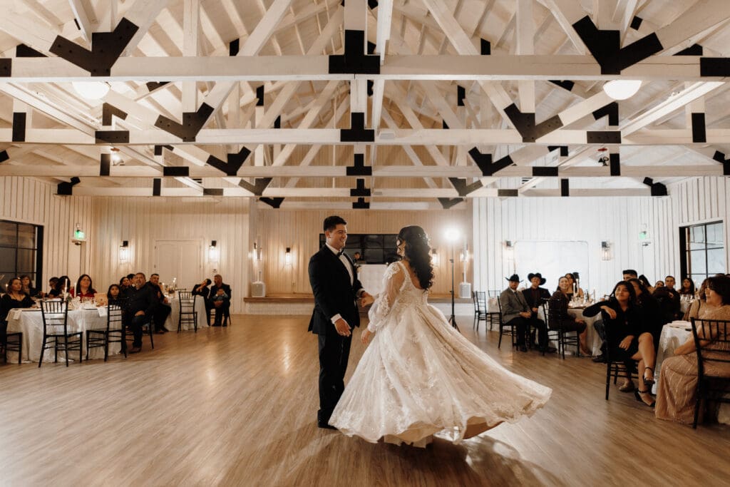 A couple is elegantly dancing in the center of a spacious, well-lit room with high, exposed wooden beams. Captured beautifully by a San Antonio wedding photographer, guests seated at tables around them watch the woman in her flowing white dress and the man in his dark suit.