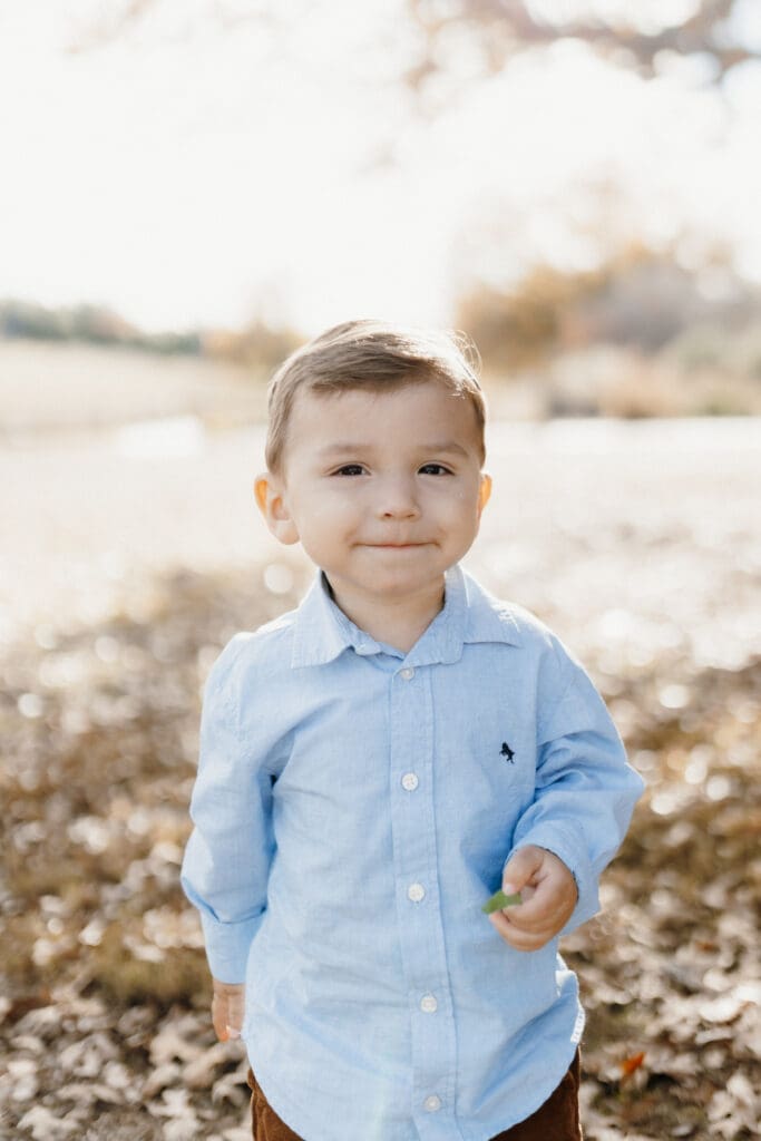A young child wearing a light blue button-up shirt and brown pants stands outdoors on a sunny day, captured impeccably by a skilled McAllen wedding photographer. The child is smiling softly, with autumn leaves scattered on the ground around them. The background of trees and a field is slightly blurred.