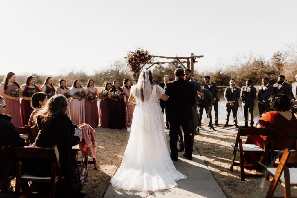 A bride in a white gown walks down an outdoor aisle with a man, approaching a groom in a suit. Bridesmaids in pink dresses and groomsmen in gray suits stand on either side. Captured by a McAllen wedding photographer, guests are seated on wooden chairs enjoying the sunny day.