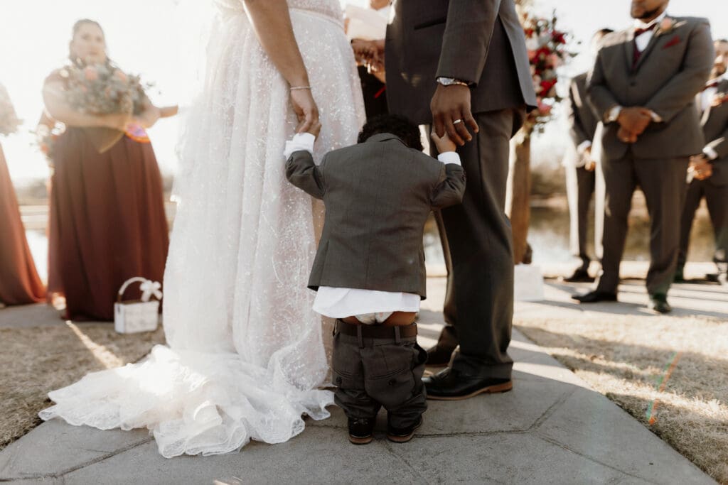 A young child in a suit, with pants slightly falling, stands between a bride in a white gown and a groom in a dark suit during an outdoor ceremony captured by a skilled McAllen wedding photographer. Bridesmaids and groomsmen are visible in the background, dressed in formal attire.