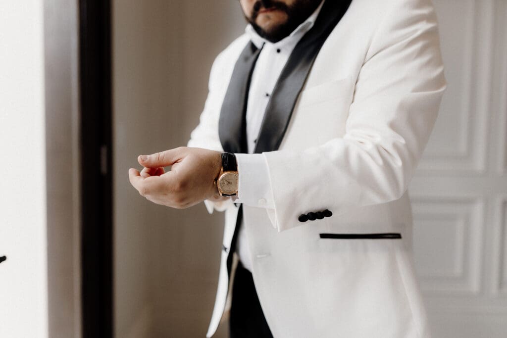 A man adjusts his cufflink, wearing a white tuxedo jacket with black accents and a black bow tie. His face is partially visible as he stands by the window, bathed in natural light—captured beautifully by a skilled wedding photographer in McAllen.