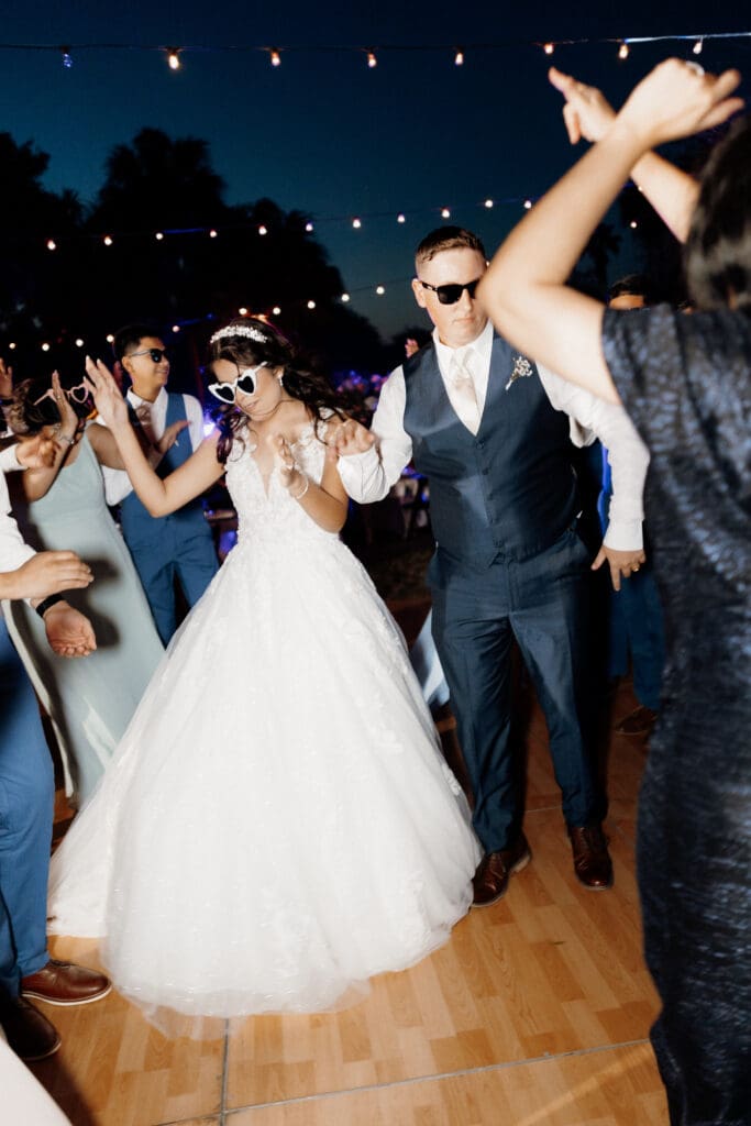 Bride and groom wearing sunglasses dance together at their beautiful outdoor wedding reception. Captured by a skilled Edinburg wedding photographer, they are surrounded by guests on a wooden dance floor under enchanting string lights. The bride dazzles in a white gown, the groom sharp in a blue suit.