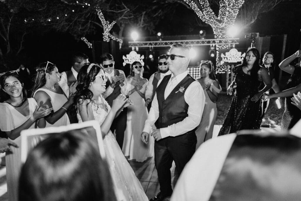A bride and groom dance at an outdoor wedding reception, surrounded by guests in formal attire. Everyone is wearing sunglasses and smiling under string lights and illuminated trees, creating a joyful and festive atmosphere. Captured by a talented McAllen wedding photographer, this scene immortalizes their special day.