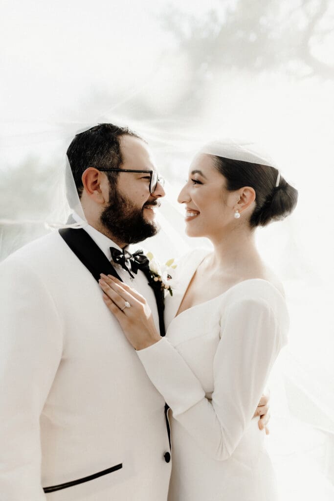 A bride and groom share a joyful moment on their wedding day, captured by a talented Edinburg wedding photographer. The bride, in a white dress and veil, smiles at the groom in glasses and a white tuxedo with a black bow tie. The background is softly blurred.