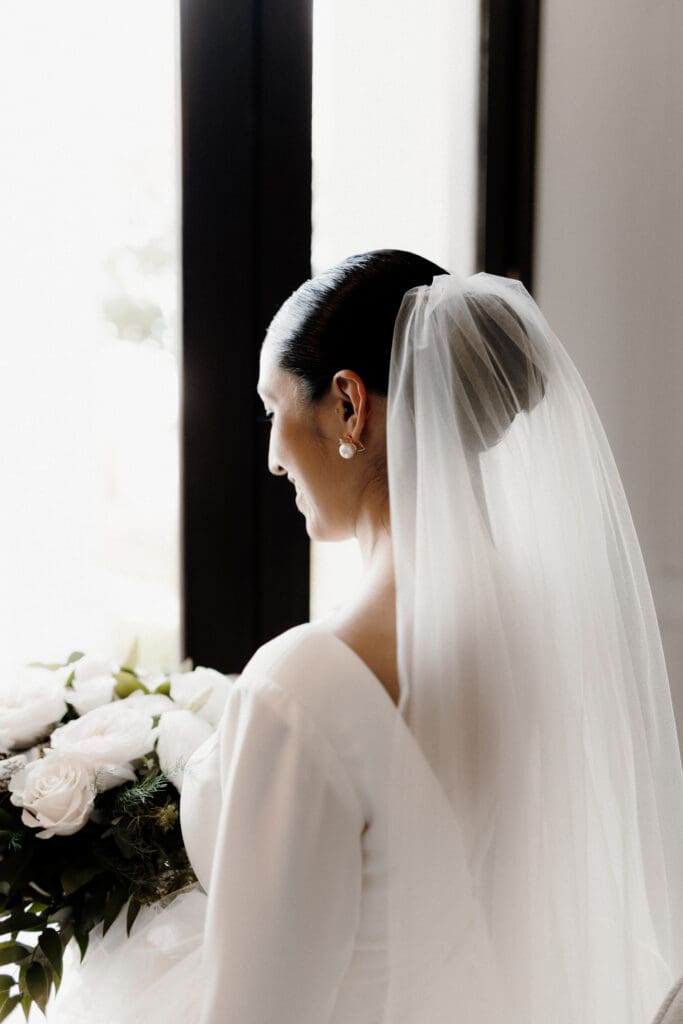 A bride in a white gown stands in front of a window, holding a bouquet of white roses and greenery. Captured by an expert San Antonio wedding photographer, her long veil drapes down her back, and her hair is styled in a bun. She gazes out the window, her face in partial profile.