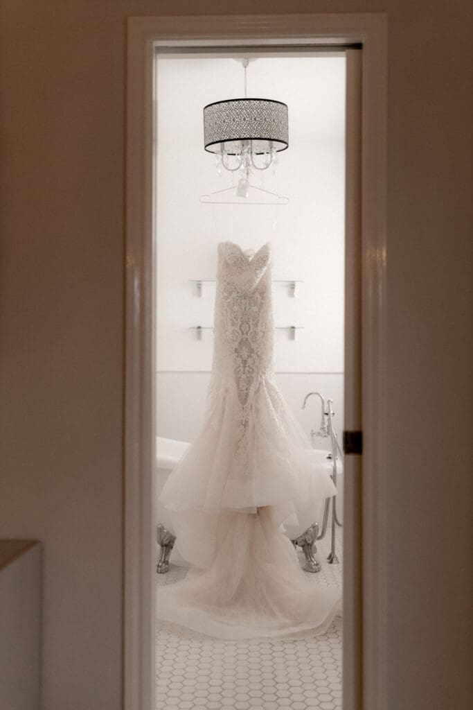 A white, strapless wedding dress with lace detail is hanging in a brightly lit bathroom, captured beautifully by a San Antonio wedding photographer. The dress features a fitted bodice and flared train, as a modern chandelier and freestanding bathtub complete the elegant scene.