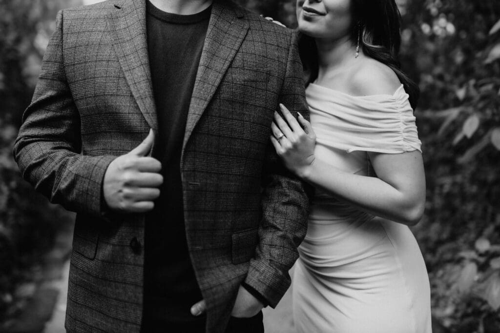A black and white image of a couple captured by a McAllen wedding photographer. The man wears a plaid jacket with his hand in his pocket, while the woman, in an off-shoulder dress, leans on his arm with her hand on his shoulder. Lush greenery is blurred in the background, adding timeless beauty.
