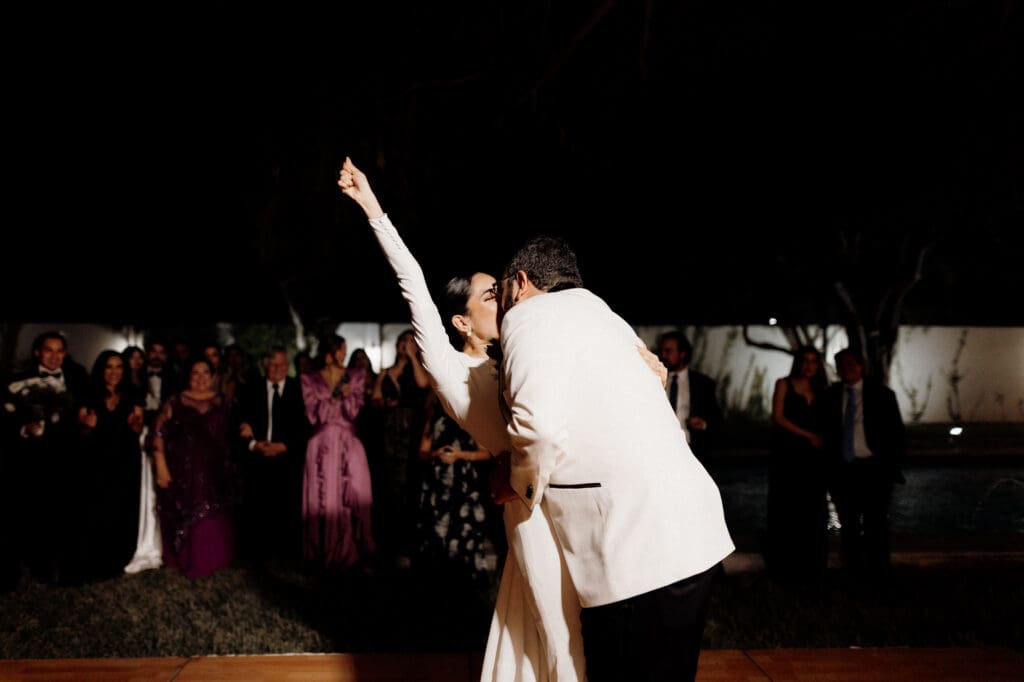 A couple dances outdoors at night, captured beautifully by an Edinburg wedding photographer. The woman in a white dress raises her arm joyfully, while the man in a suit leans in close. Guests in formal attire watch the dance, with trees and a lit background.