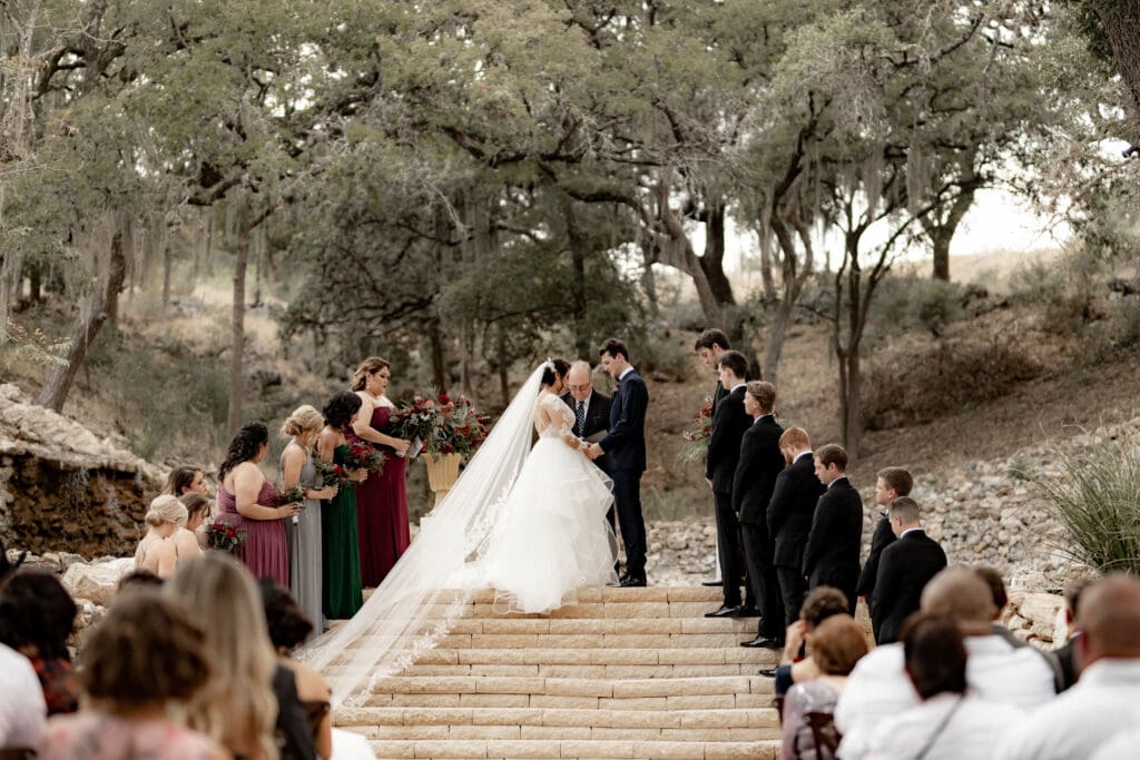 A couple stands on outdoor steps exchanging vows at their enchanting ceremony. The bride, radiant in a white gown with a flowing veil, and the groom, dashing in a dark suit. Bridesmaids in colorful dresses and groomsmen nearby complete the picturesque scene captured by a San Antonio wedding photographer amid tall trees.