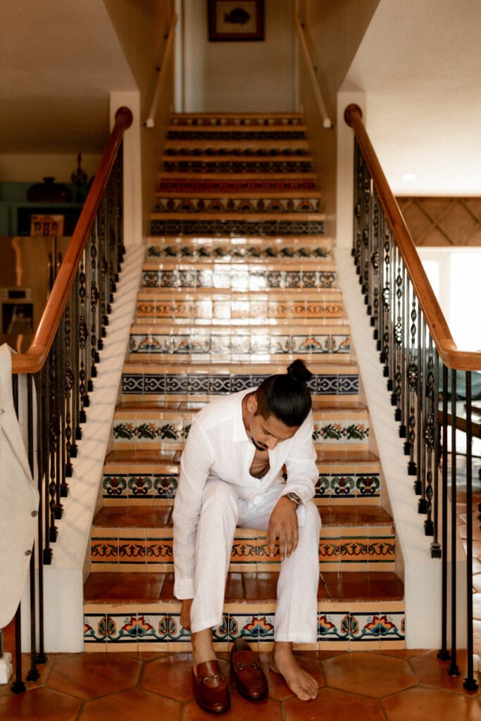 A person with a bun, dressed in a white shirt and pants, sits on the colorful tiled stairs of a warmly lit room, putting on brown loafers. The settings wrought iron railings and wooden accents hint at the artful eye of a San Antonio wedding photographer.