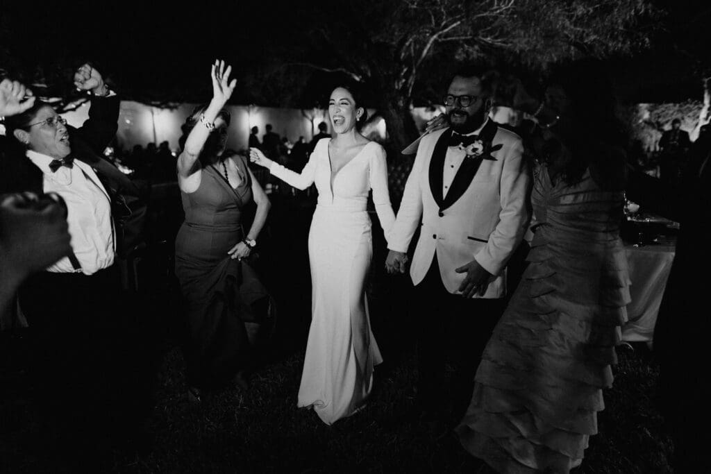 A joyful couple in formal attire dances with guests at an outdoor wedding reception, expertly captured by a McAllen wedding photographer. The bride wears a long dress, and the groom dons a tuxedo. Lively expressions and celebratory gestures fill the scene, beautifully rendered in black and white.