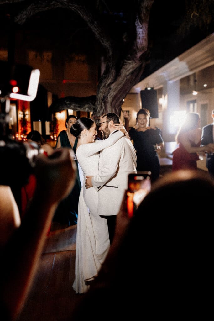 A couple is embracing and dancing closely under soft lighting at an indoor event, captured beautifully by a San Antonio wedding photographer. The woman wears a white dress, and the man dons a light-colored suit, while guests in the dimly lit background snap photos with their phones.