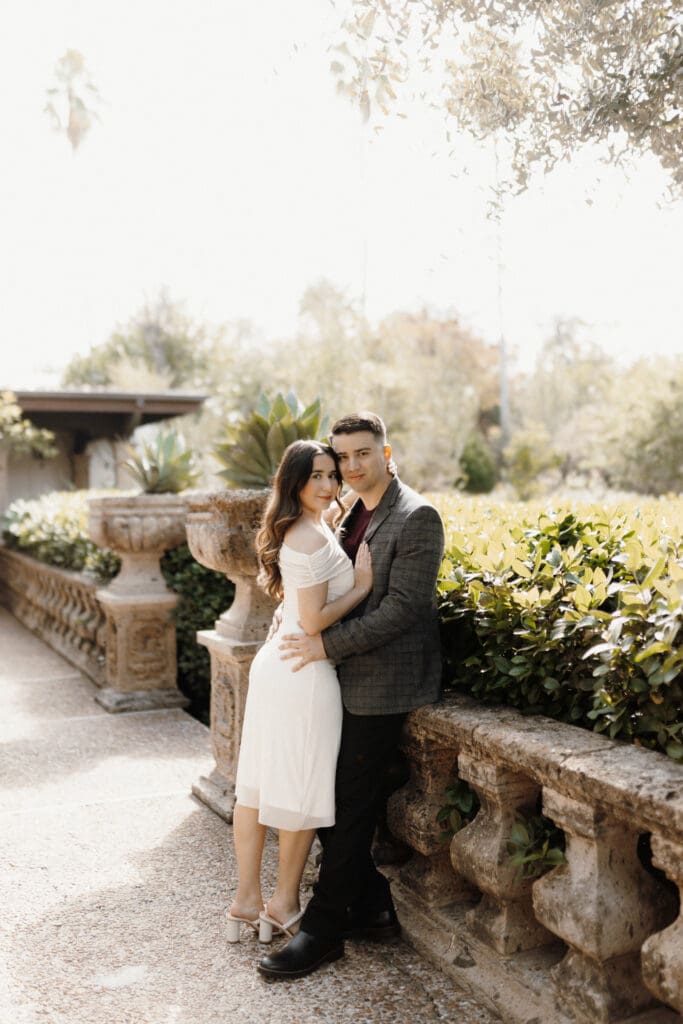 A couple stands closely together, posing on a sunlit stone walkway lined with greenery. The woman wears a white dress and the man dons a checkered blazer. Captured by a talented wedding photographer from McAllen, they are surrounded by lush foliage and an ornate stone railing.