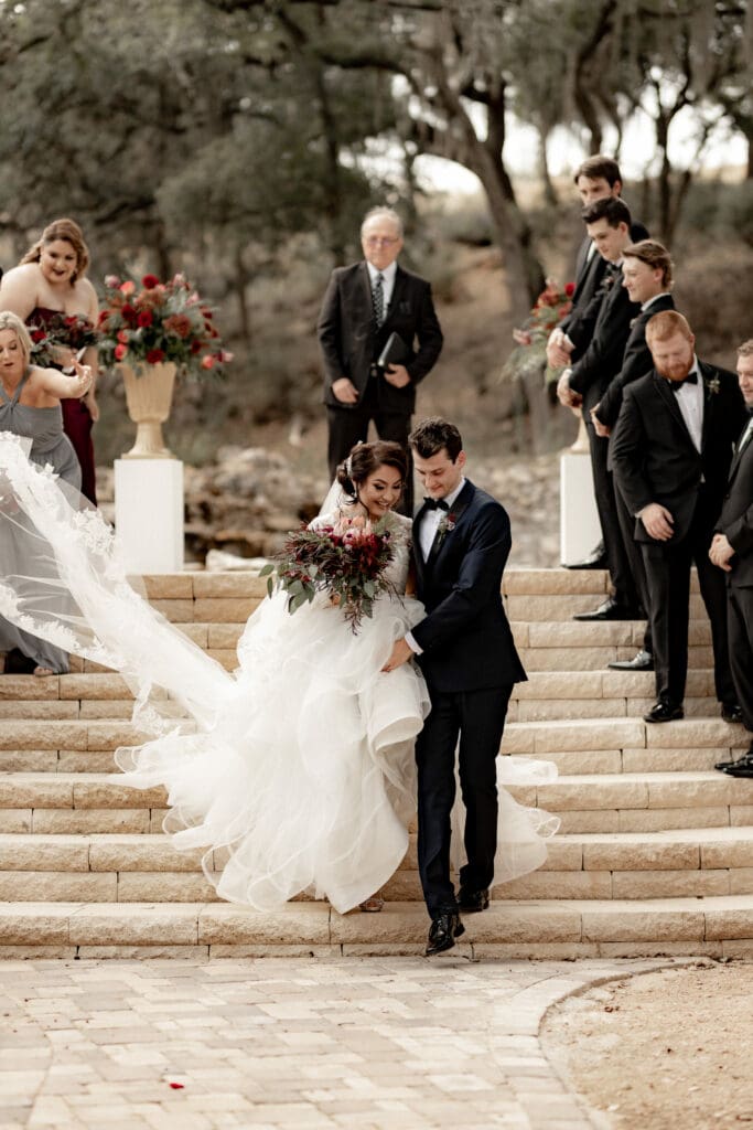 A bride and groom descend stone steps, smiling and holding hands as a San Antonio wedding photographer captures the moment. The bride wears a voluminous white gown, while the groom dons a dark suit. Wedding party and guests stand nearby, holding floral arrangements against a backdrop of trees and greenery.