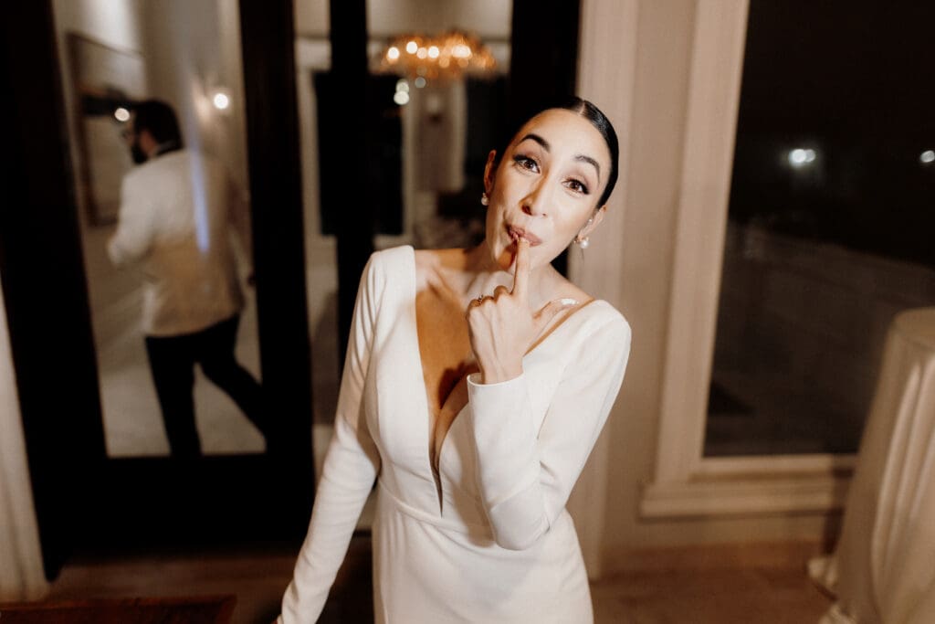 A person in an elegant white outfit playfully poses with one finger near their lips. Captured by a McAllen wedding photographer, a large mirror and chandelier adorn the background, along with a blurred figure in a suit.