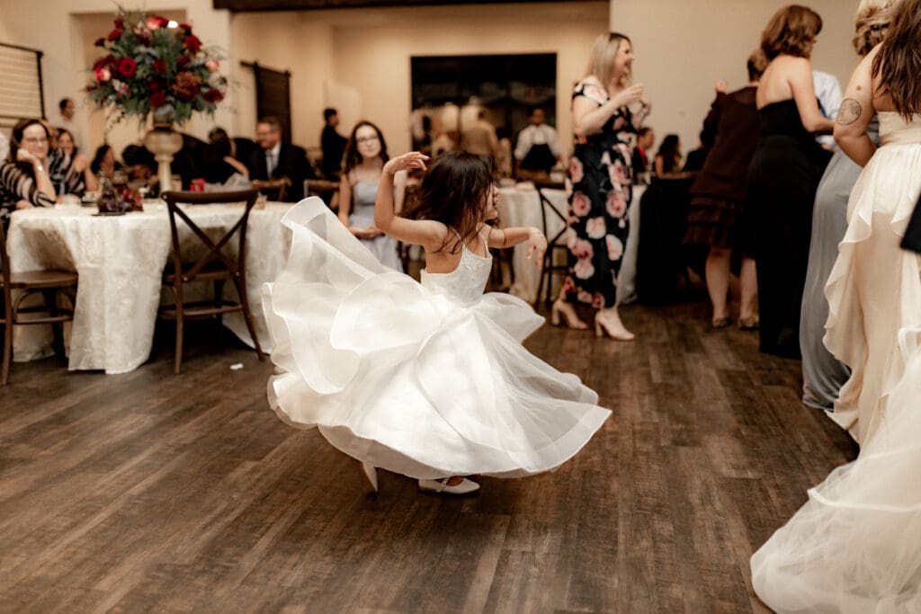 A young girl in a flowing white dress twirls joyfully on a wooden dance floor at a party, captured beautifully by a wedding photographer in McAllen. Guests sit at round tables with white tablecloths and floral centerpieces, while others stand and watch in the warmly lit room.