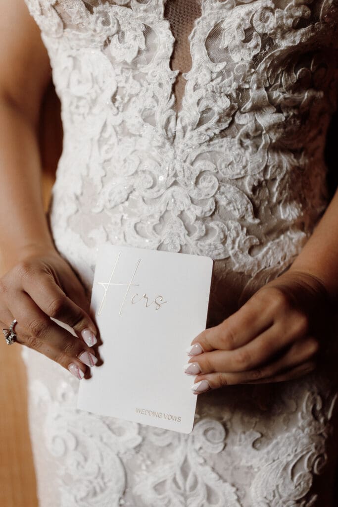 A bride in a lace wedding dress holds a white booklet titled Hers with Wedding Vows at the bottom. Her nails are manicured, and she wears a ring on her left hand, captured beautifully by a talented Edinburg wedding photographer.