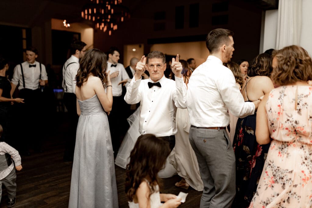 A group of people dancing at a formal event, captured by a talented San Antonio wedding photographer. A man in a white shirt and black bow tie stands in the center, pointing playfully. Elegant dresses and suits adorn the attendees, with soft lighting and chandeliers adding to the ambiance.
