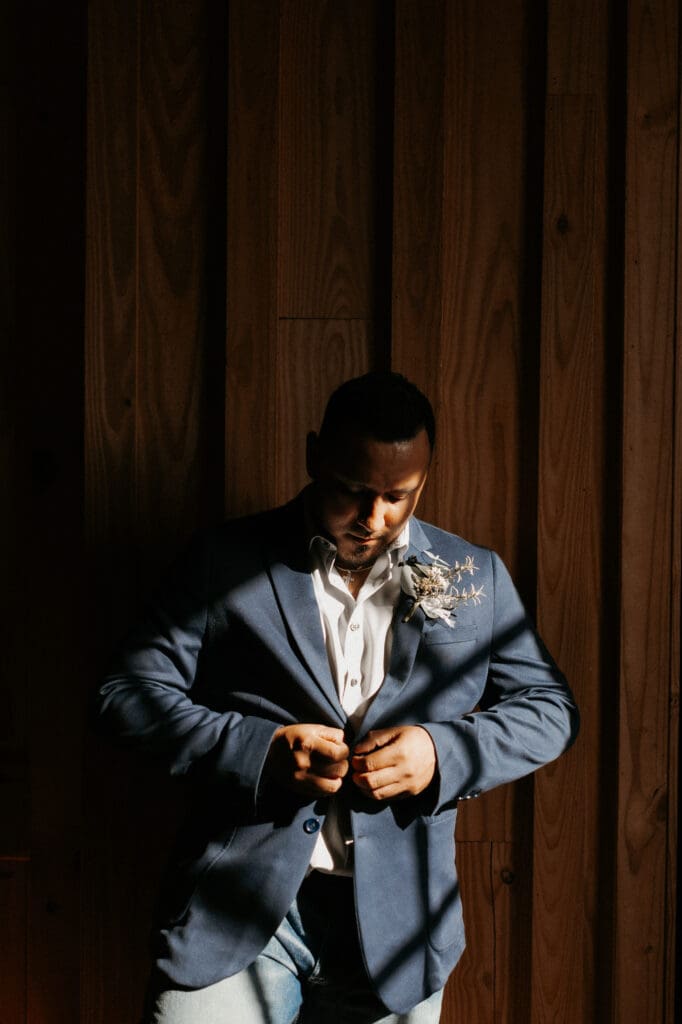 A man in a dark blue suit with a light shirt stands against a wooden wall, buttoning his jacket. As the sunlight casts dramatic shadows, highlighting his focused expression, he appears ready for the lens of an Edinburgh wedding photographer. A small boutonniere decorates his lapel.