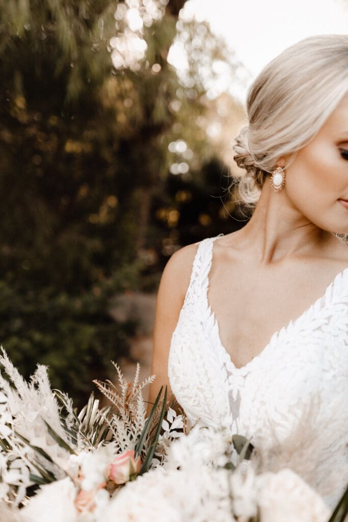 Bride in a lace wedding gown, holding a bouquet of flowers. Her hair is styled in an elegant updo, and she wears pearl earrings. Captured by a McAllen wedding photographer, the soft, natural background suggests an enchanting outdoor setting.