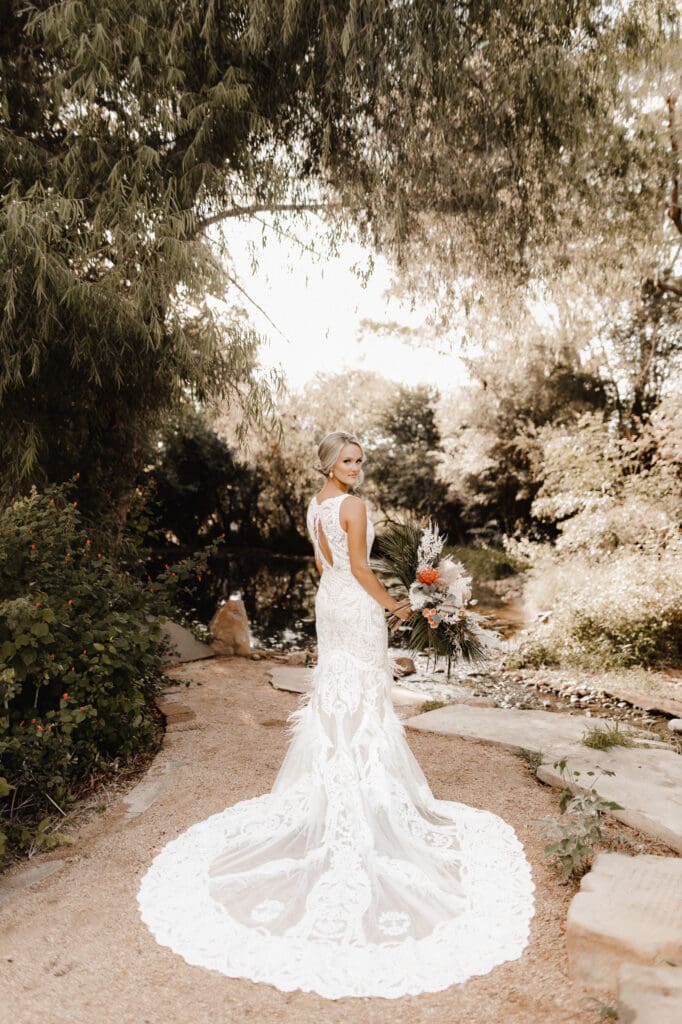 A bride in a lace wedding dress stands on a garden path, holding a bouquet. Her dress has a long, flowing train. Captured by an Edinburg wedding photographer, she is surrounded by lush greenery and trees, with a small pond in the background.