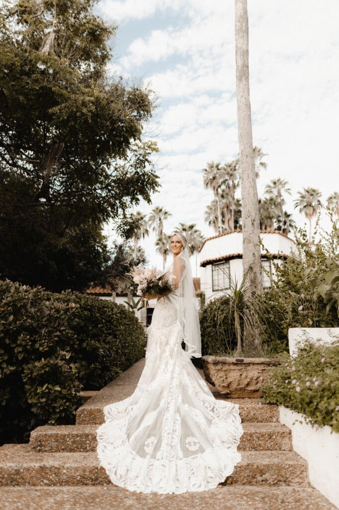 A bride in a lace wedding gown stands on stone steps, enveloped by lush greenery. Holding a bouquet, she glances over her shoulder as her long train cascades behind her. The partly cloudy sky and tall palm trees complete the scene, beautifully captured by an Edinburg wedding photographer.