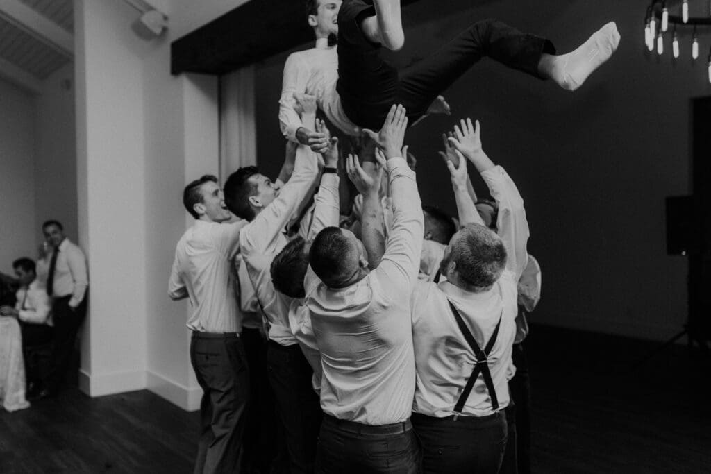 A group of people in formal attire joyfully lifts a person in the air during a celebration indoors. Captured by an Edinburg wedding photographer, the black-and-white image conveys the dynamic movement and excitement of the scene.