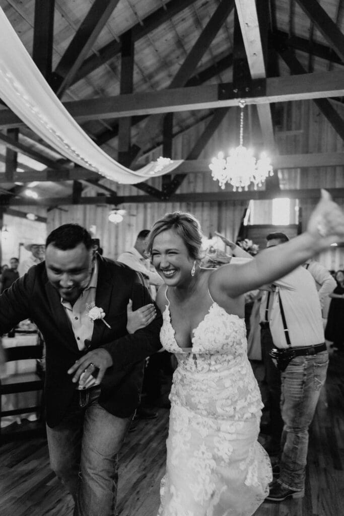 A bride in a lace gown and groom in a dark suit joyfully dance under wooden beams and chandeliers. They smile broadly, captured brilliantly by a wedding photographer in McAllen, surrounded by guests in the warmly lit reception hall.