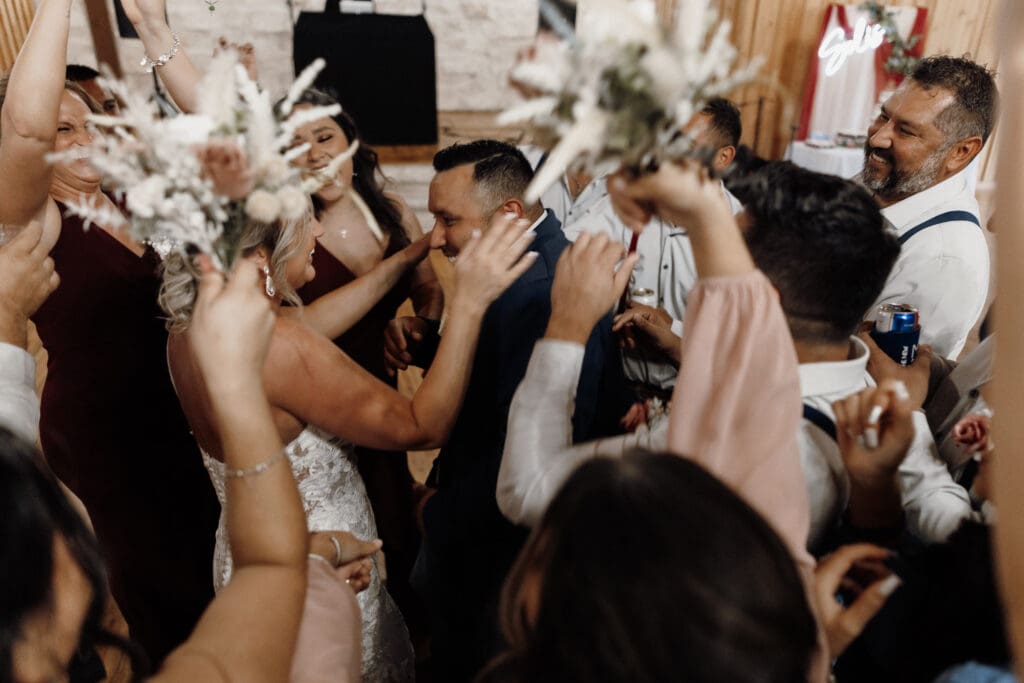A bride and groom dance surrounded by joyful guests, expertly captured by a McAllen wedding photographer. The bride holds her bouquet as everyone raises their arms in celebration. The atmosphere is lively and festive, with people close together in a warmly lit room.