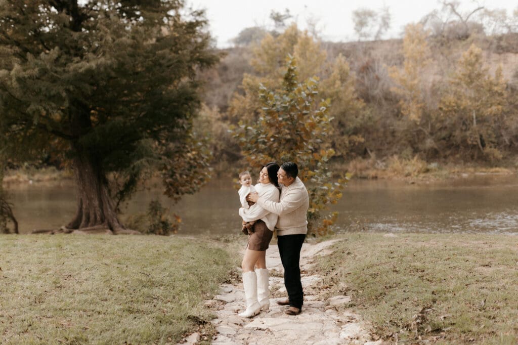 A couple stands on a stone path beside a river with autumn trees in the background, captured perfectly by a McAllen wedding photographer. Holding their young child, all dressed in warm clothing, the grassy area and serene water enhance the peaceful atmosphere.