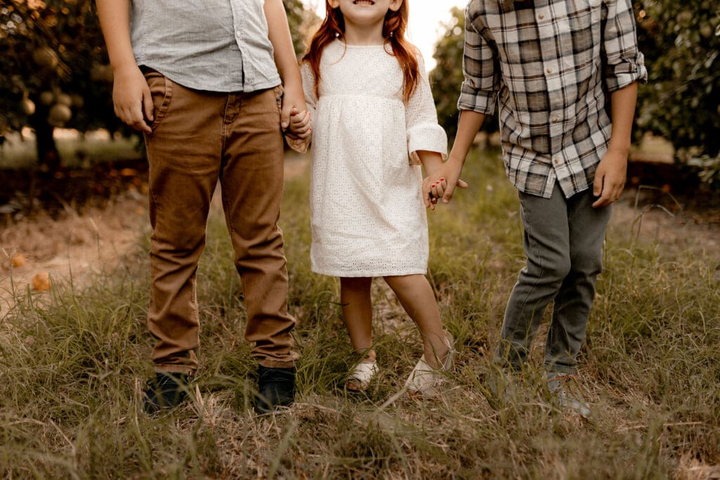 Three children stand holding hands in a grassy field, captured beautifully by an Edinburg wedding photographer. The child on the left wears a light shirt and brown pants, the child in the middle dons a white dress, while the child on the right sports a plaid shirt with gray pants.