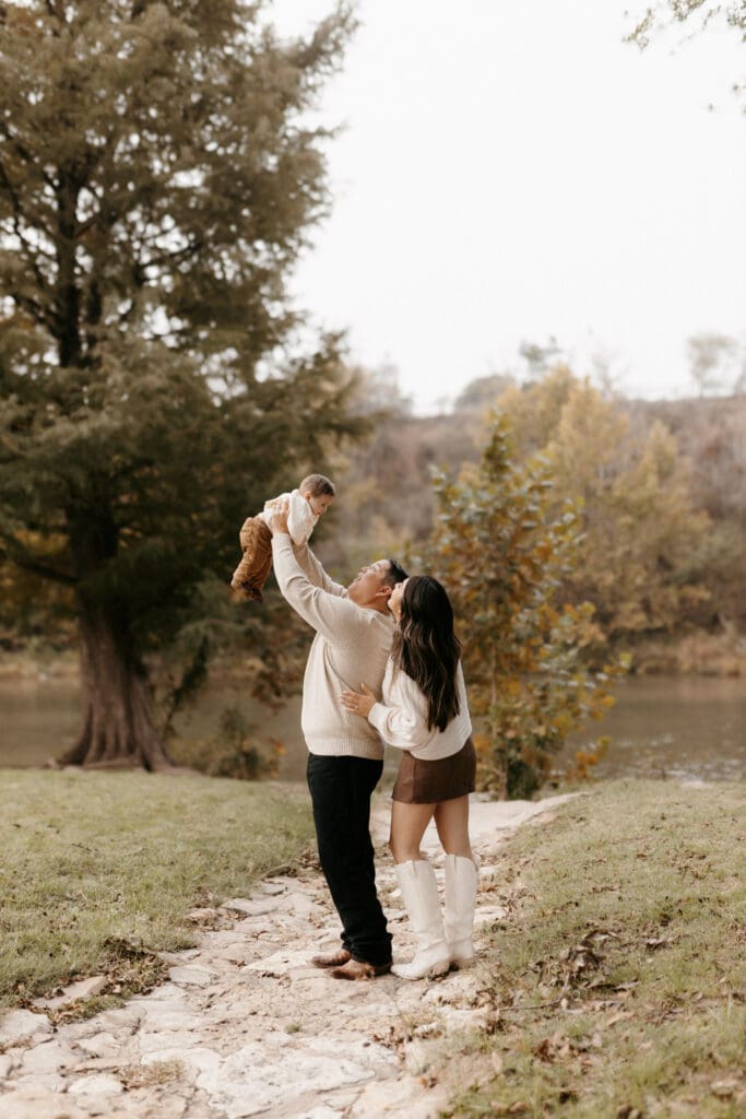 A couple stands on a stone path near a riverbank, with trees in the background. The man joyfully lifts a baby into the air while the woman, wearing a cream sweater and boots, looks on lovingly with her arm around him. Captured by an Edinburg wedding photographer, the scene is serene and nature-filled.