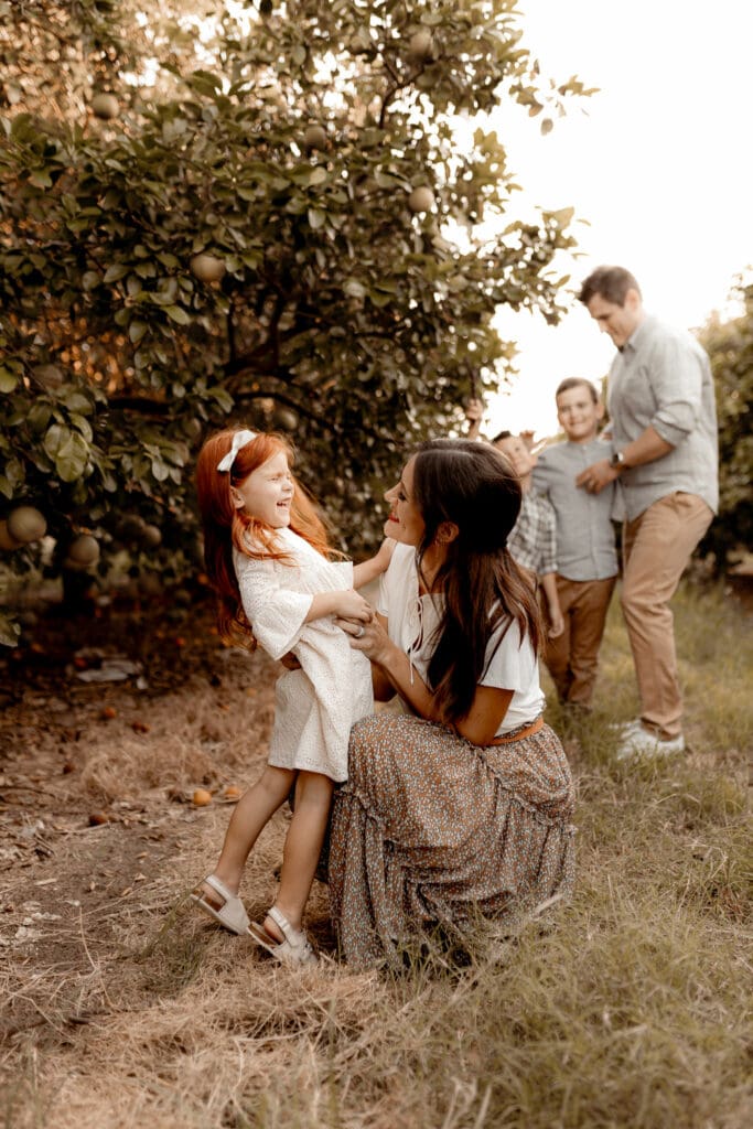 A woman crouches and smiles at a young girl in a white dress next to an orange tree, captured beautifully by a McAllen wedding photographer. In the background, two adults and another child stand, smiling. The scene is outdoors, and the mood is joyful and relaxed.