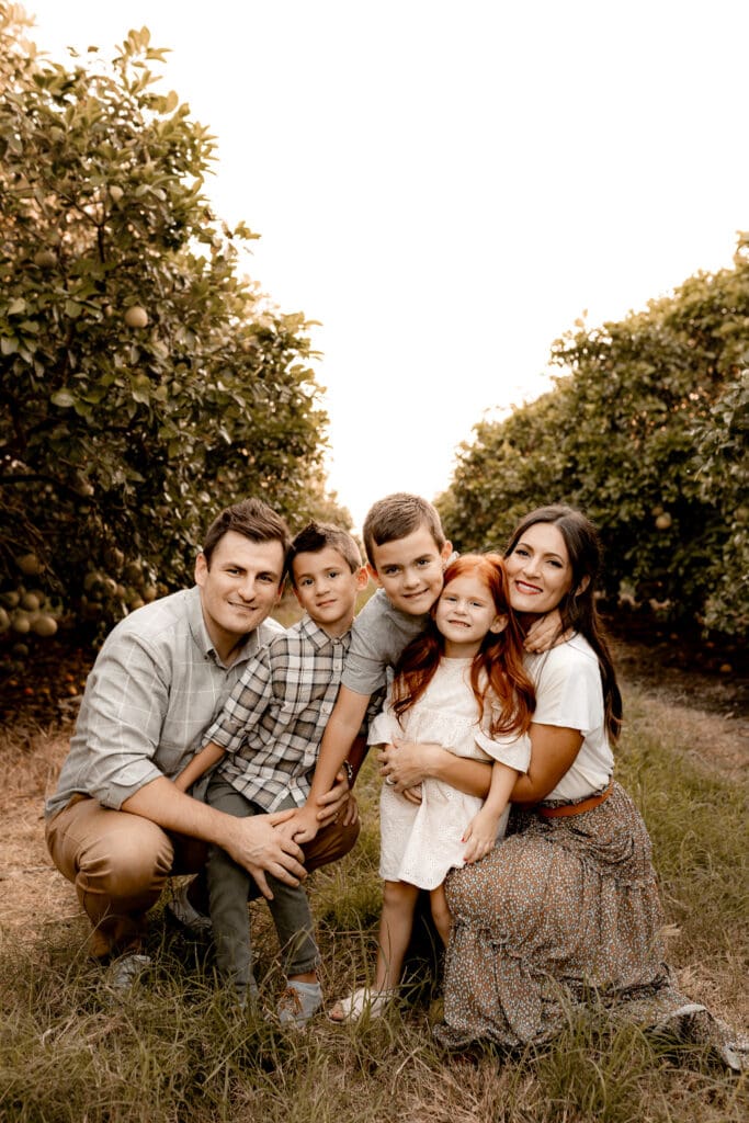 A family of five poses together in an orchard, kneeling on grass between rows of trees. The man and boy on the left wear plaid shirts, while the woman wears a white top. The girl with long red hair in a white dress smiles warmly—a perfect moment captured by an Edinburg wedding photographer.