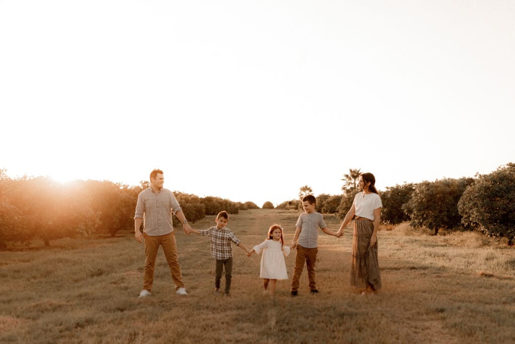 A family of five holds hands while standing in a sunlit orchard, captured beautifully by an Edinburg wedding photographer. The group includes two adults and three children as the setting sun casts a warm glow across the grassy field, with trees and the sunset painting the perfect backdrop.