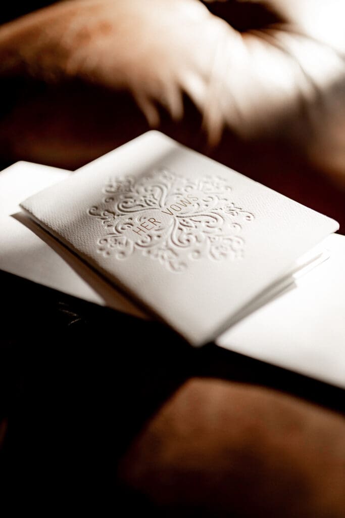 Close-up of a textured, embossed wedding vow booklet on a soft, fabric background. The cream-colored booklet bears the words HER VOWS visible in the design. Captured by an Edinburg wedding photographer, the soft, warm lighting creates a gentle glow on the surface.