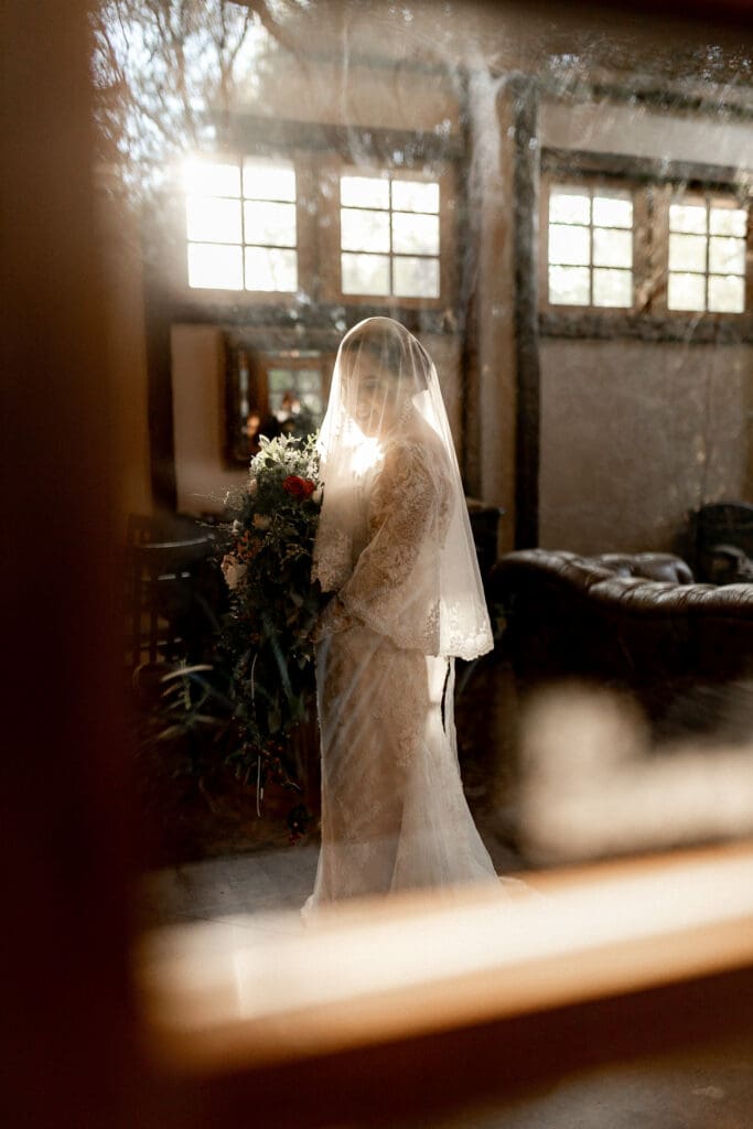 Bride in a lace gown and veil stands indoors holding a large bouquet, softly illuminated by sunlight streaming through high windows. Captured by a skilled San Antonio wedding photographer, the scene is viewed through a pane of glass, adding a dreamy, intimate ambiance.