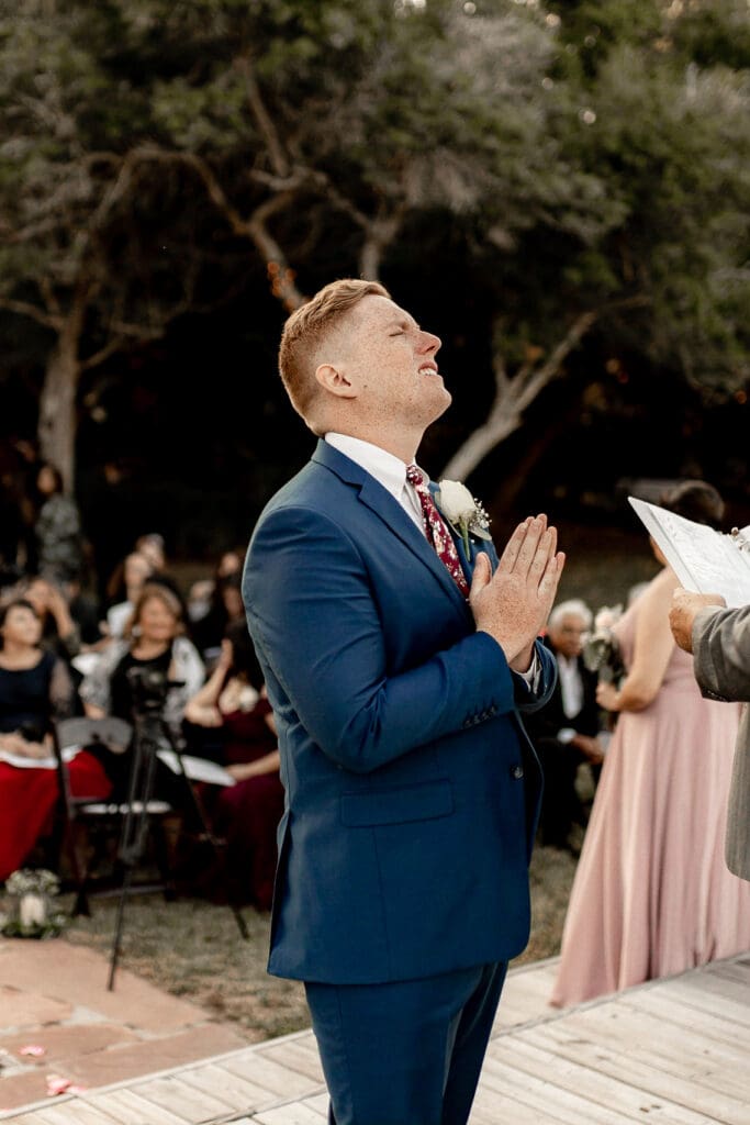 A person in a blue suit stands outdoors at a wedding ceremony on South Padre Island, looking upward with hands clasped. They are next to an officiant holding papers, as the wedding photographer captures the moment. Seated guests and trees create a picturesque background.