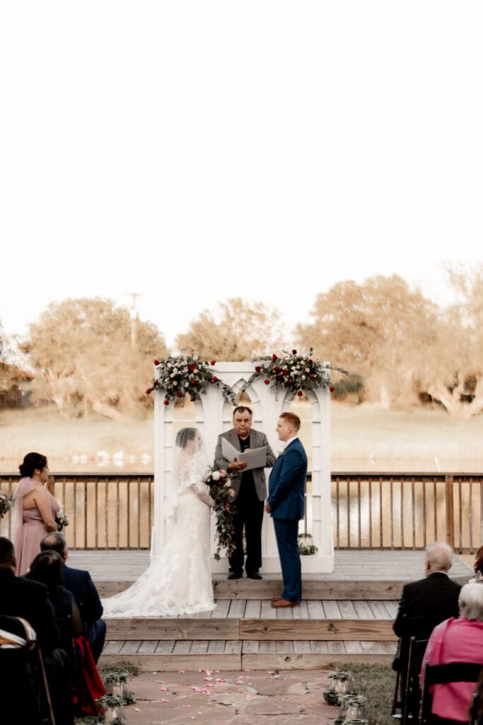 A bride and groom stand at an outdoor wedding ceremony on a wooden deck with a floral arch behind them. Captured by an Edinburg wedding photographer, the officiant stands in the center, book in hand. Guests are seated on either side, with trees and a field setting the perfect backdrop.