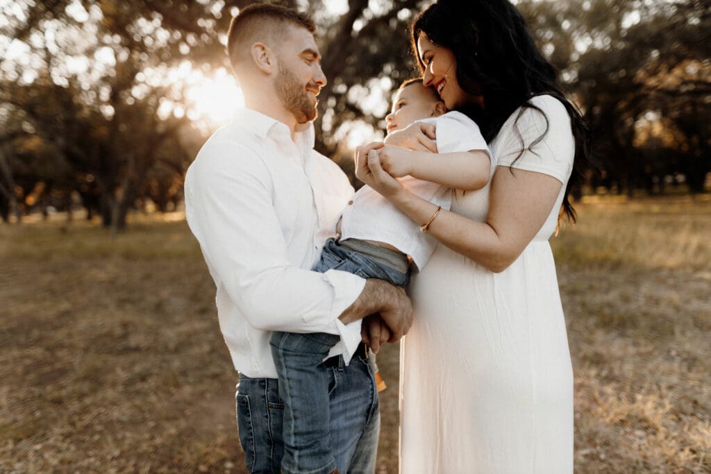 A couple stands in a sunlit field, captured beautifully by a skilled Edinburg wedding photographer. Smiling joyfully at each other and their young child, the woman in a white dress holds the child close while the man, in a white shirt, supports them on his arm. Trees provide a serene backdrop.